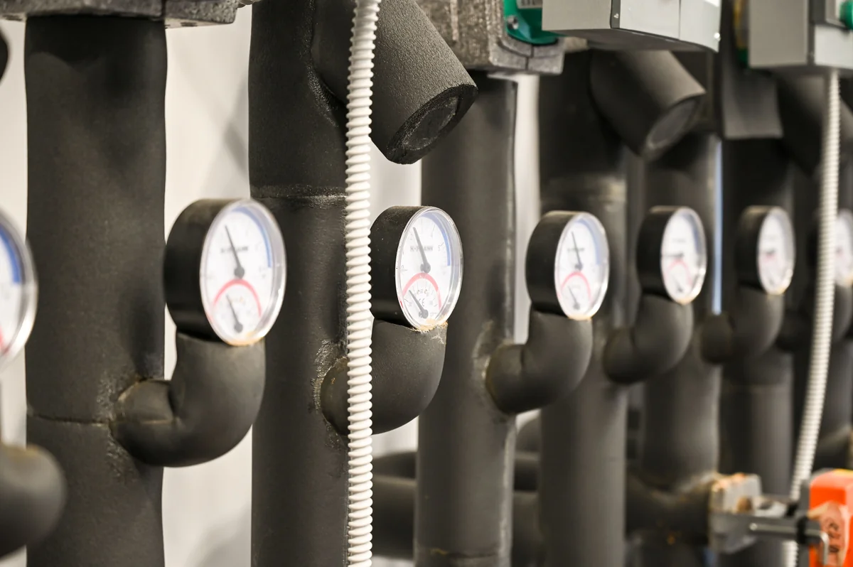 Landscape photograph of repeated dark insulated industrial pipes with round pressure gauges and connecting conduits; no people are visible.