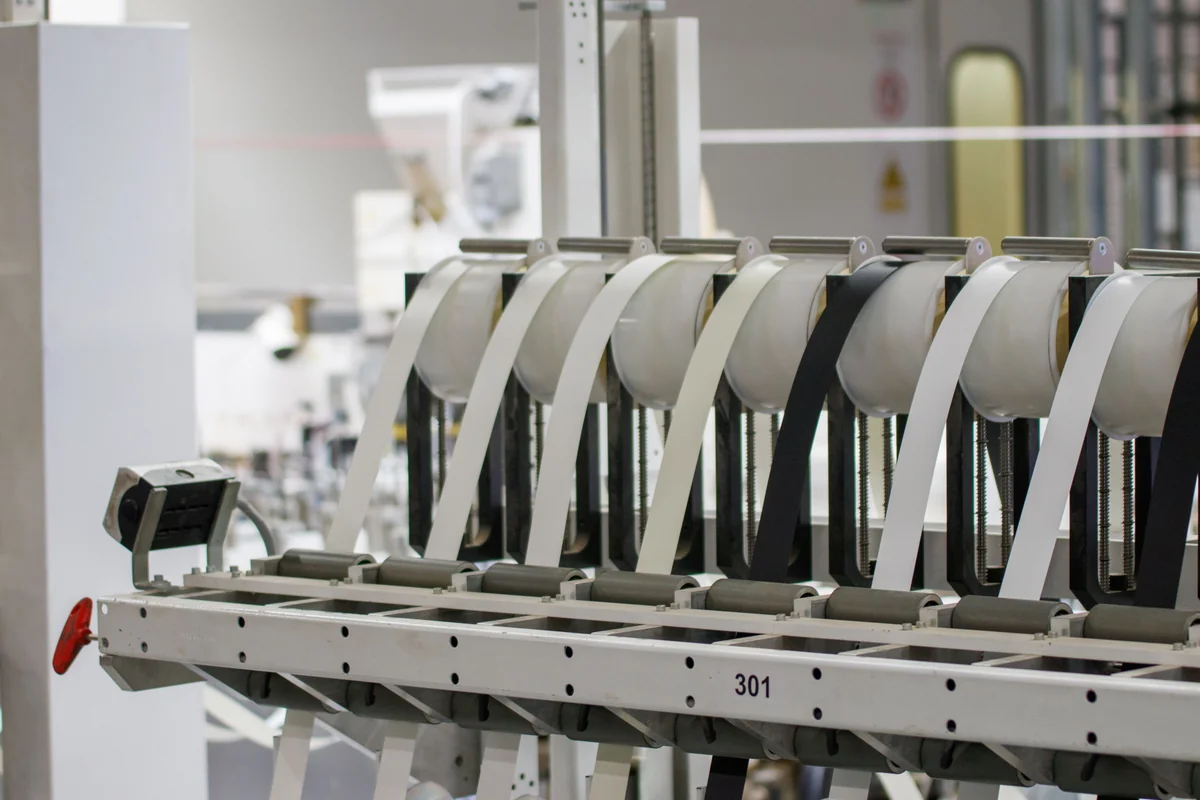White and black material strips feed through parallel rollers in a clean industrial manufacturing machine.