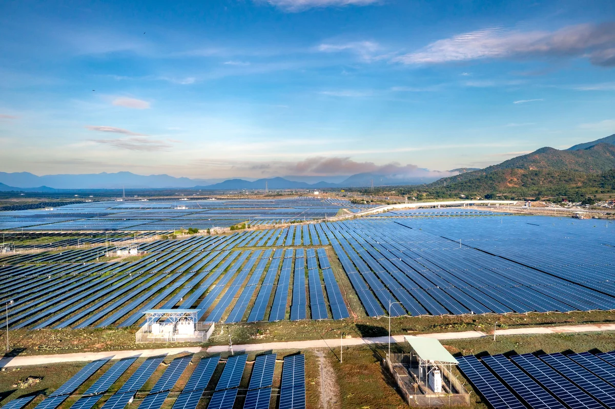 Aerial landscape showing rows of dark solar panels across open land with distant mountains and sky.