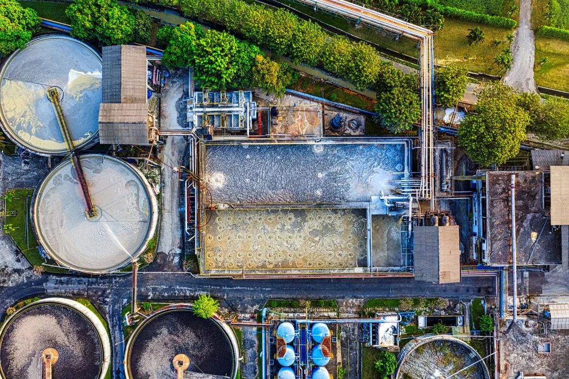 Aerial view of circular treatment tanks, rectangular basins, buildings, and pipework at a wastewater treatment facility surrounded by trees.