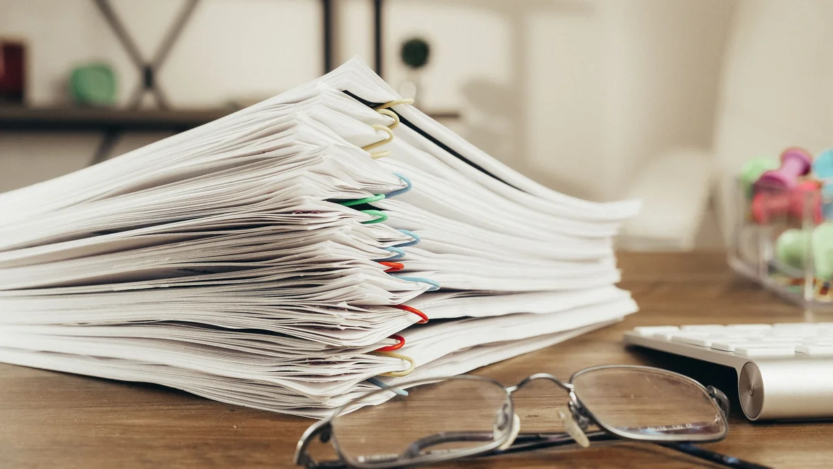 Stacks of clipped paper documents on a wooden desk with eyeglasses and a blurred neutral office background.