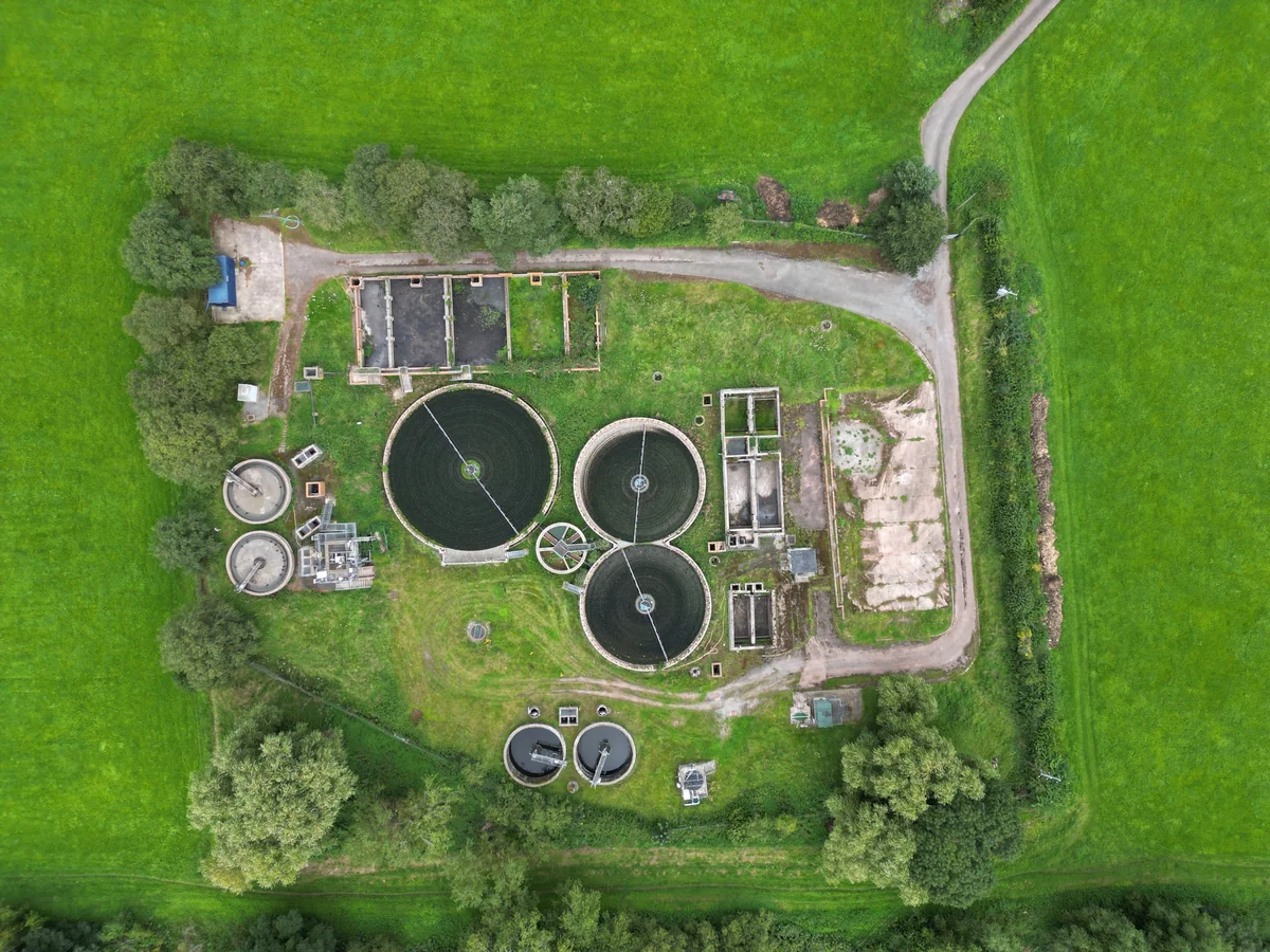 Aerial view of a wastewater treatment plant with circular settling tanks, rectangular basins, access roads and trees in green fields.