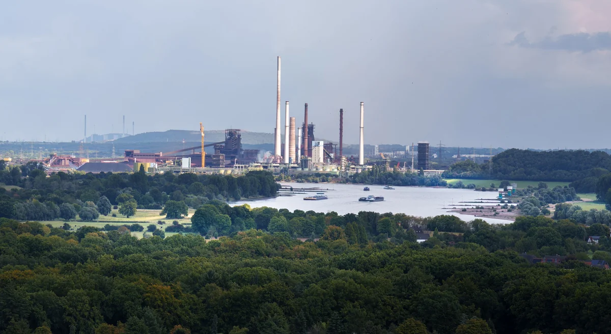 Factory complex with smokestacks and industrial structures beyond a band of leafy trees, with a river in the middle distance.