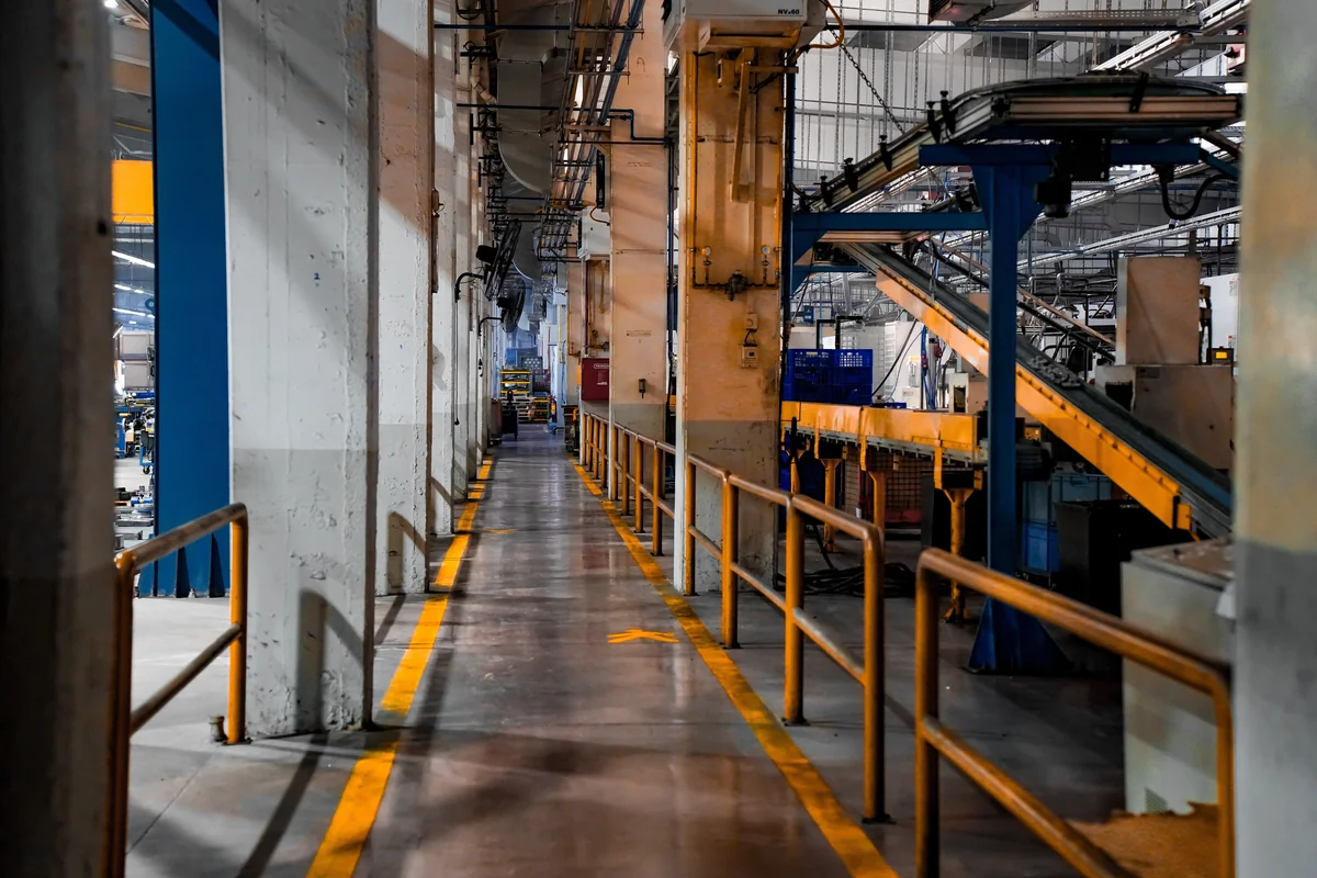 A wide interior view of a manufacturing facility with conveyor machinery, overhead structures, and an empty marked walkway.