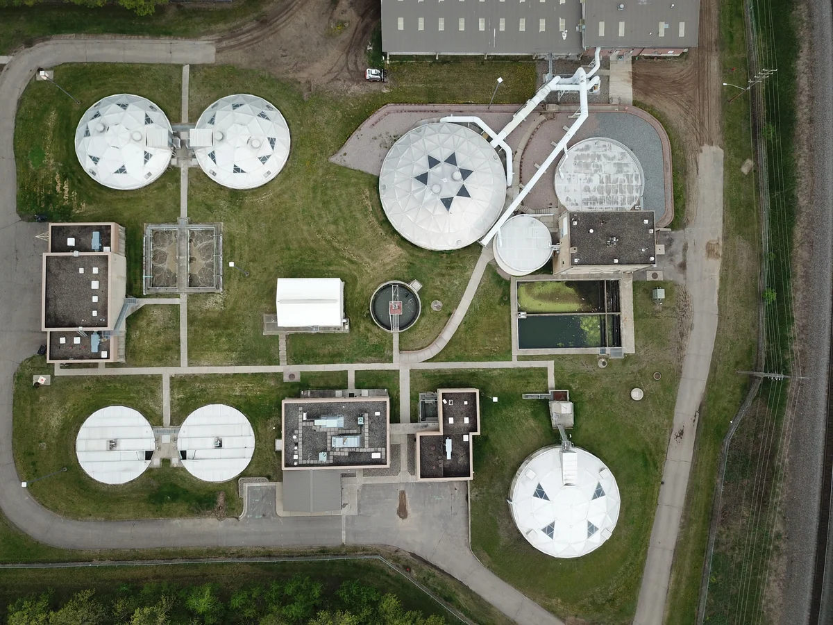 Aerial view of a water treatment facility with circular covered tanks, rectangular basins, connected pipes, buildings, roads and grass.