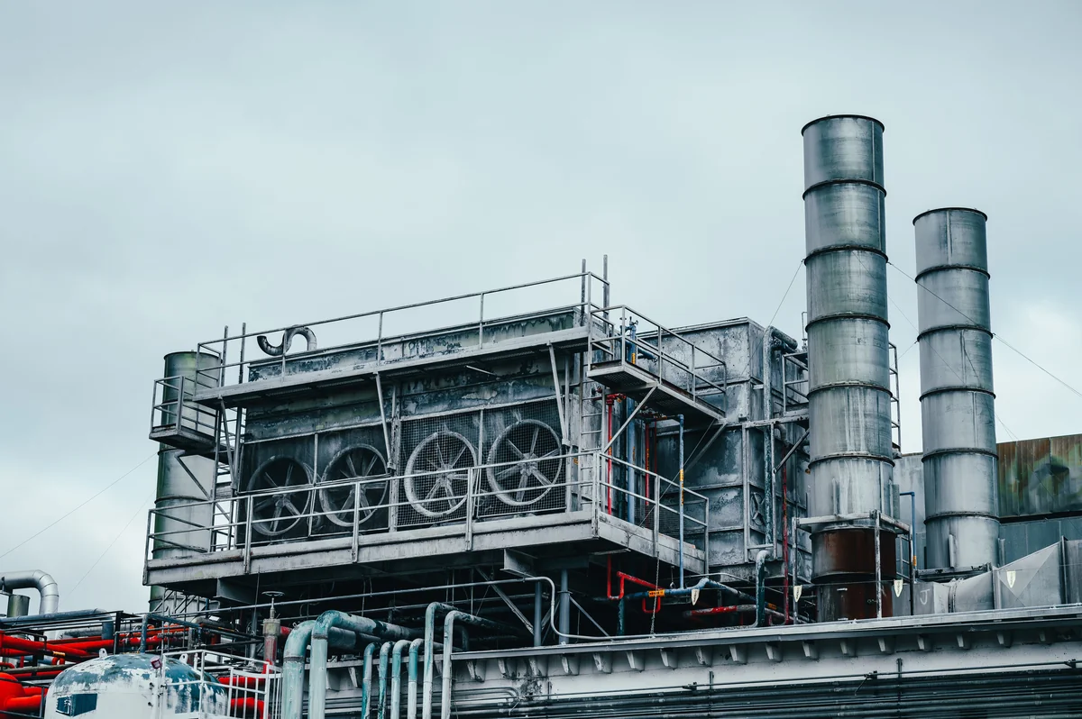 Exterior of an industrial facility with metal chimneys, large ventilation fans, railings, tanks and interconnected pipes beneath a cloudy sky.