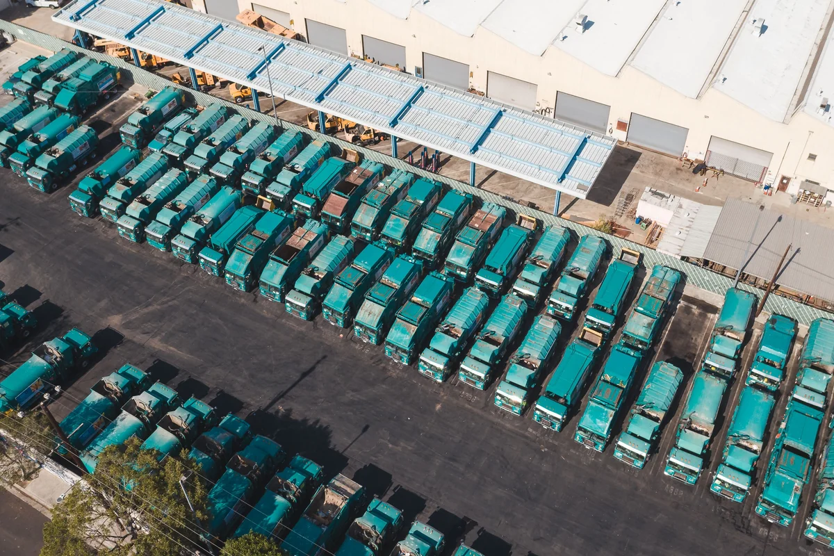 Aerial landscape view of rows of turquoise transport trucks parked beside a large industrial warehouse.