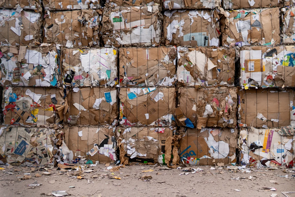 Large bales of recycled cardboard stacked outdoors in rows, with loose paper and cardboard on the ground.