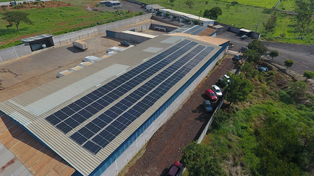 Aerial view of an unbranded industrial building with rows of solar panels on its roof, surrounded by paved yards, trees, and green fields.