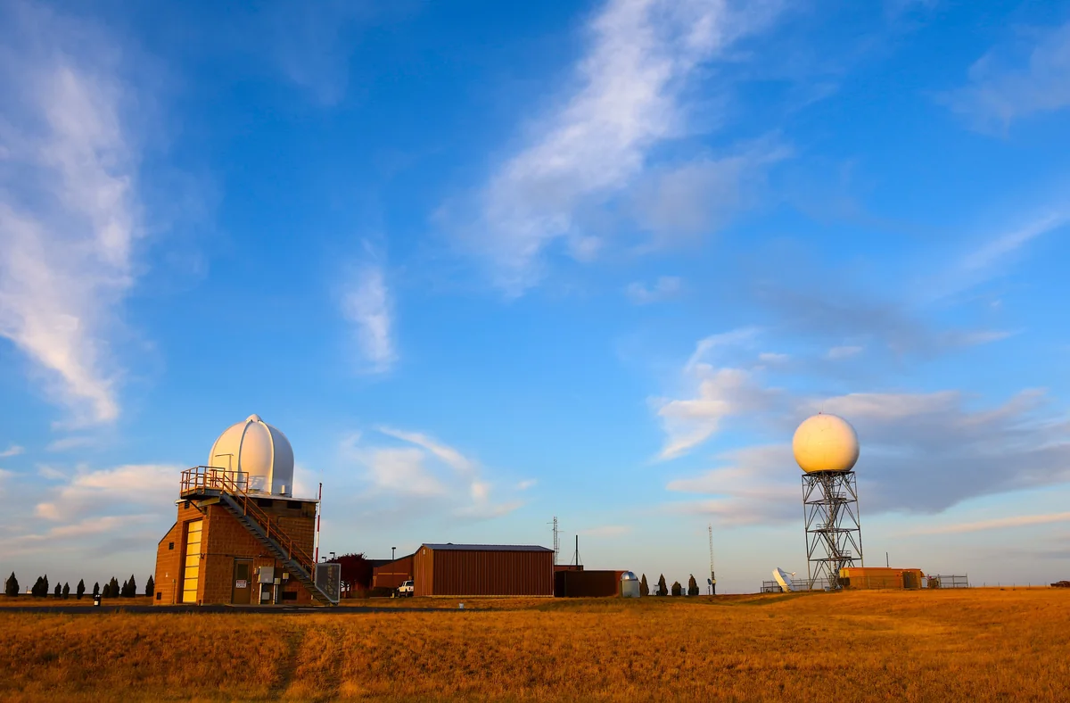 Landscape photograph of an unoccupied weather radar station with two radar domes and monitoring instruments in an open field beneath a blue sky with scattered clouds.