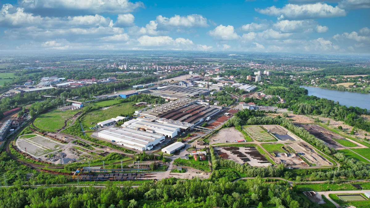 Aerial landscape view of a large industrial complex surrounded by green fields, trees and roads under a bright sky.