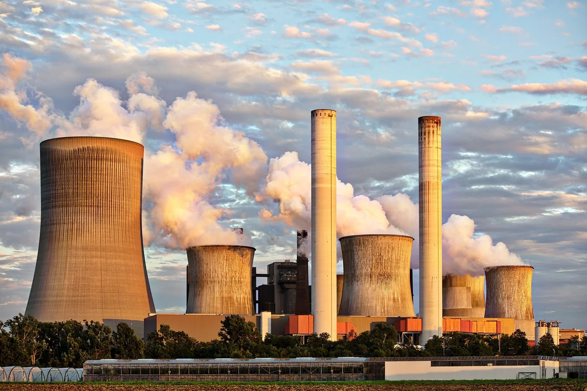 Power plant cooling towers and chimneys release white steam above a tree-lined industrial site under a partly cloudy sky.