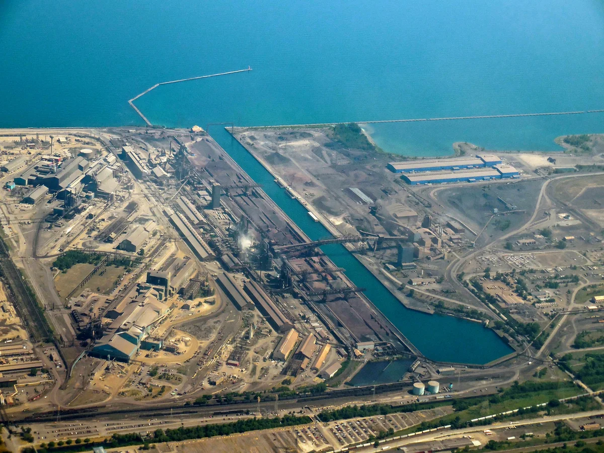 Aerial view of a large steel manufacturing complex with rail lines, processing buildings, storage areas, roads, a canal and adjacent blue water.