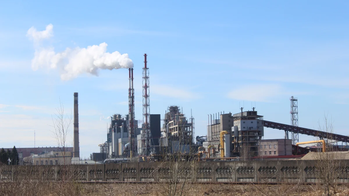 Unbranded industrial manufacturing plant with processing structures, several tall chimneys, pipework and white emissions against a clear blue sky.