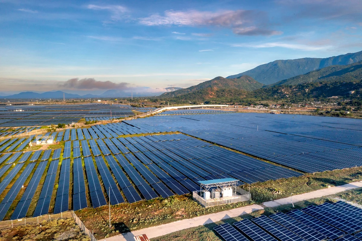 Aerial landscape view of extensive rows of solar panels across open fields, with distant mountains and a clear sky.