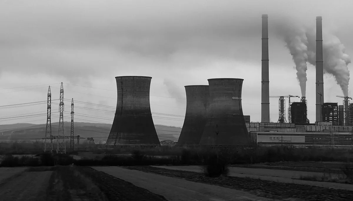 Black-and-white landscape view of cooling towers, tall chimneys, power lines and industrial buildings under a cloudy sky.