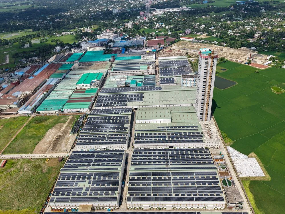 Aerial landscape view of a sprawling industrial complex with solar-panel-covered roofs, surrounding green fields, roads, and no visible people.