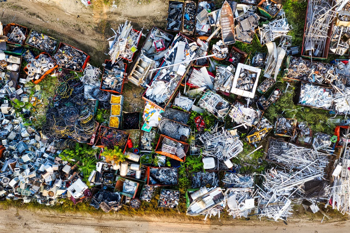 Aerial landscape photograph of many separated scrap-metal piles and containers arranged across an outdoor recycling yard, with dirt access lanes and patches of vegetation.