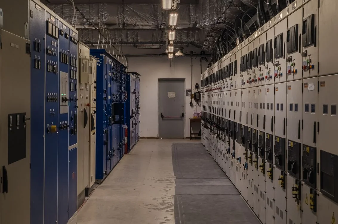 Empty industrial control room with rows of electrical panels, wiring and overhead lights.