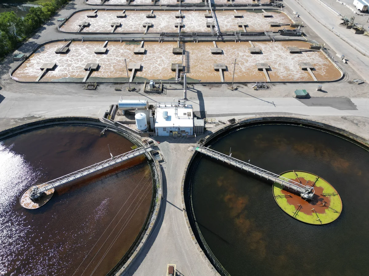 Aerial view of a water treatment plant with large circular tanks, rectangular basins, access roads and surrounding vegetation.