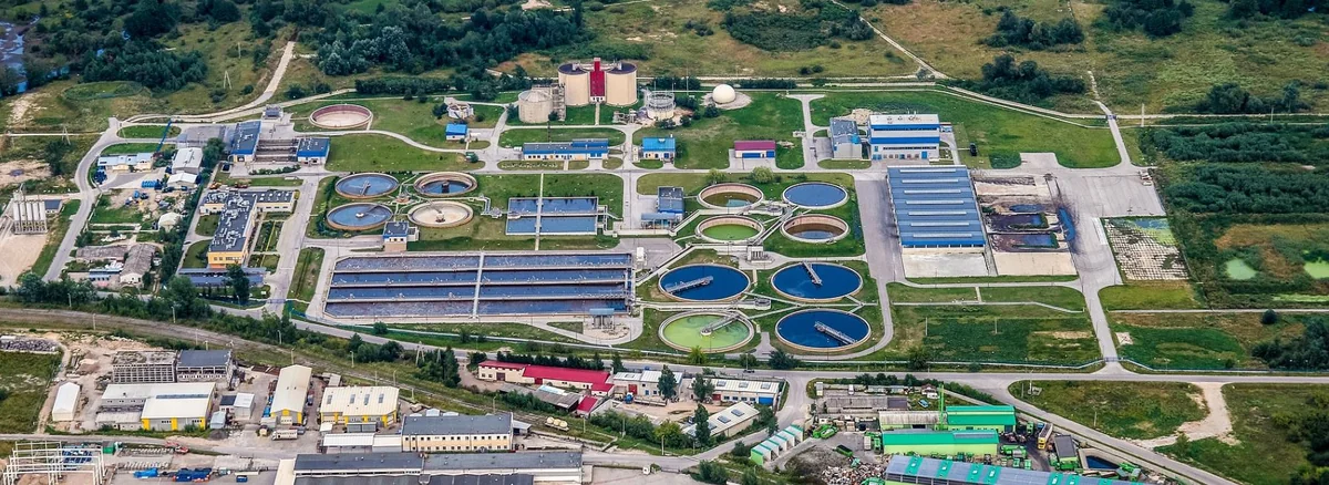 Aerial landscape view of a wastewater treatment facility with circular clarifier tanks, rectangular treatment basins, service buildings, access roads and surrounding greenery.