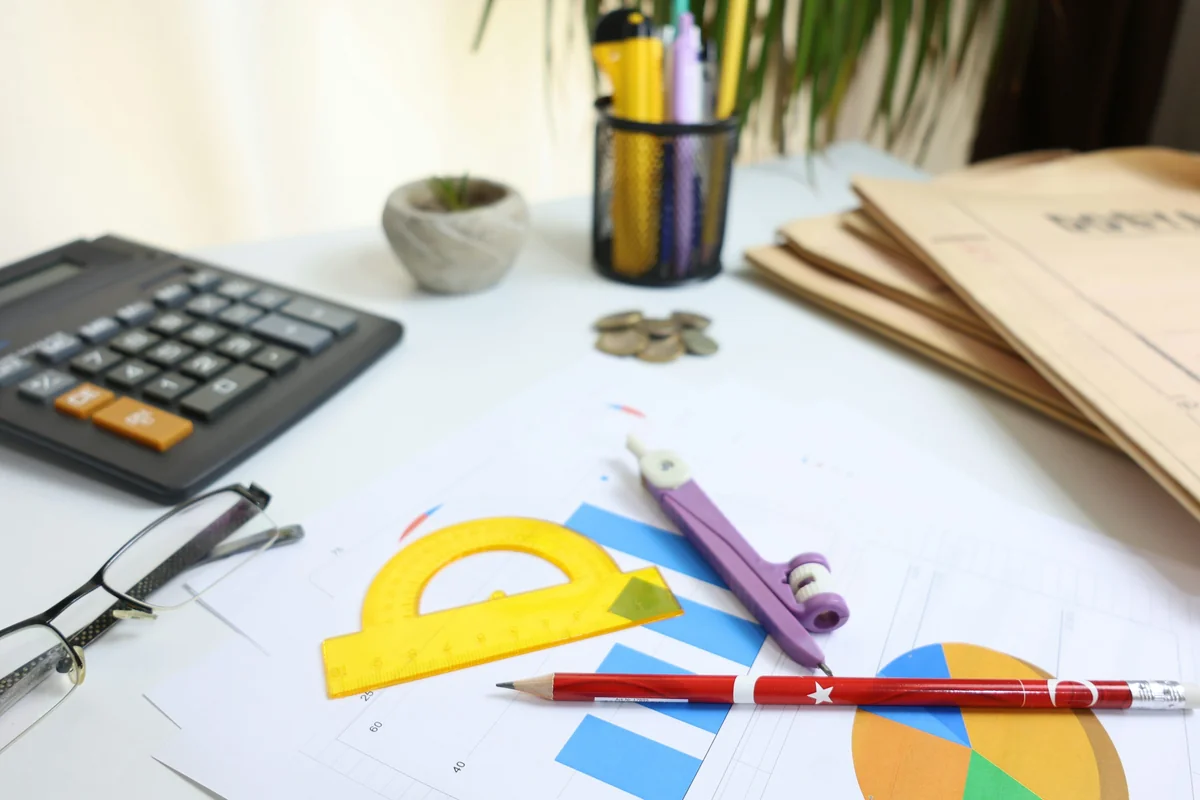 Landscape photograph of an unoccupied desk with a calculator, notebook, pencil, eyeglasses and a small potted plant; no readable data or people are visible.