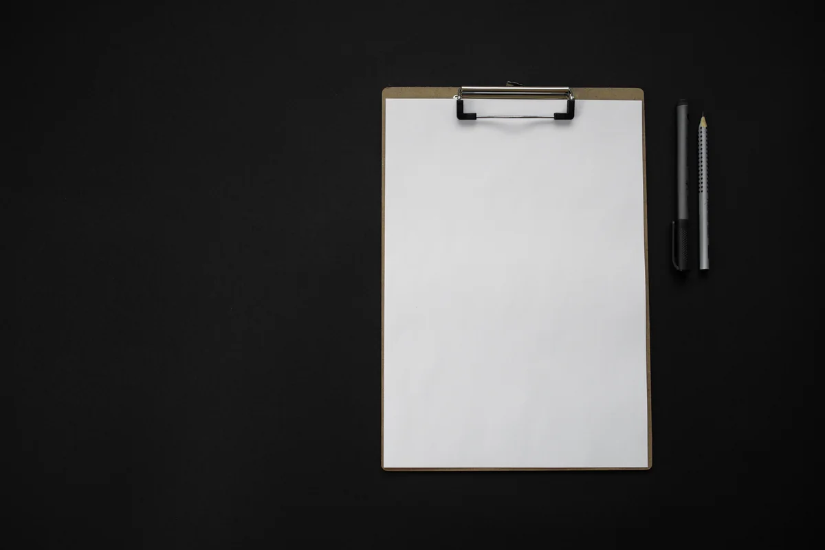 Flat-lay photograph of a blank white sheet clipped to a dark clipboard, with a pen and pencil on a black background.