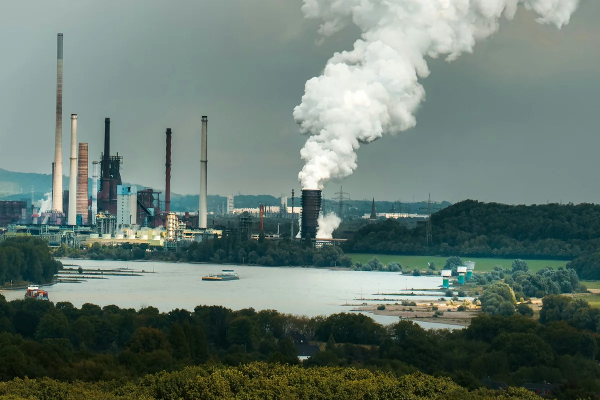 Industrial factory buildings and smokestacks across a river, with wooded greenery and a cloudy sky.