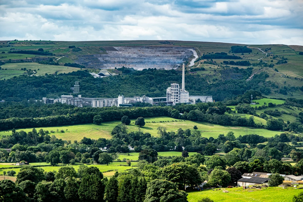 A distant cement factory among green hills and fields under a cloudy sky.
