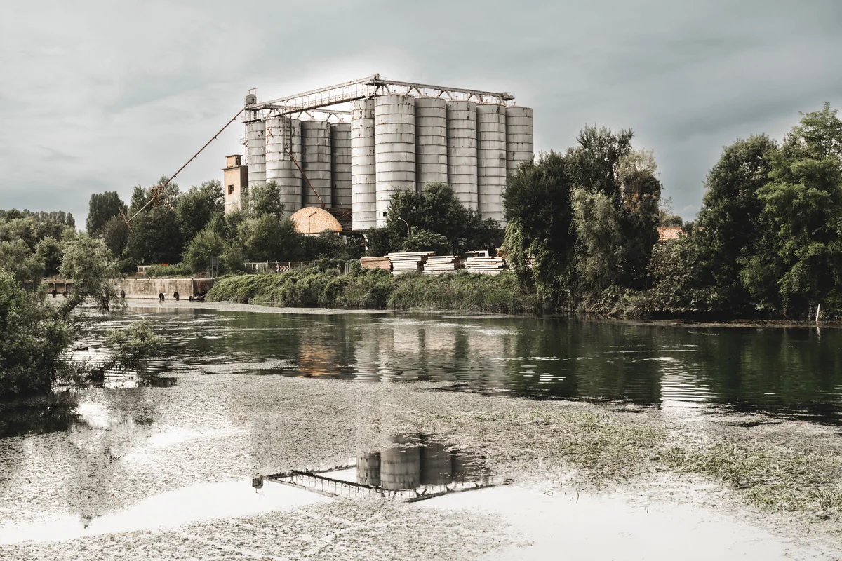 Large industrial silos beside a tranquil river, with lush riverside trees and reflections in the water under a cloudy sky.