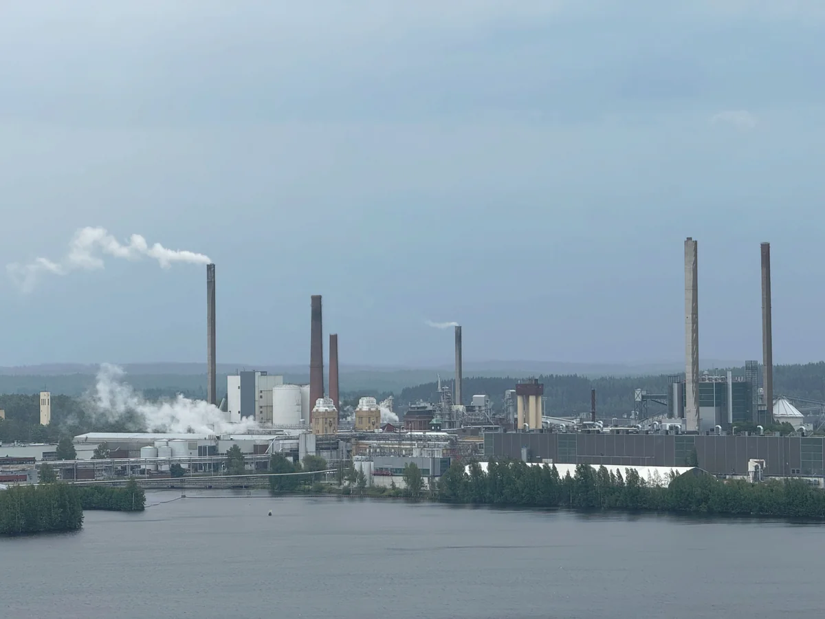 Factory complex with multiple smokestacks and white steam plumes across a waterfront, backed by forested hills beneath a cloudy sky.