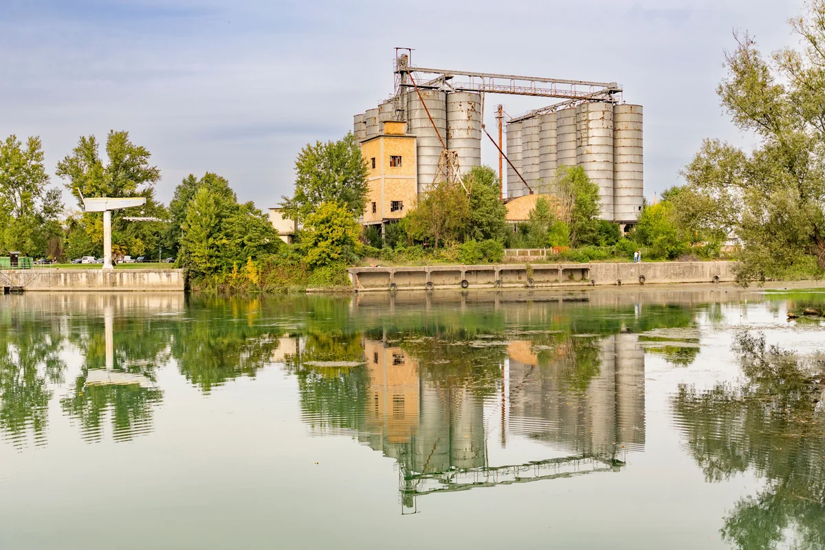 Industrial silos and a low riverside facility stand among leafy trees beside calm water, with the structures reflected on the surface.