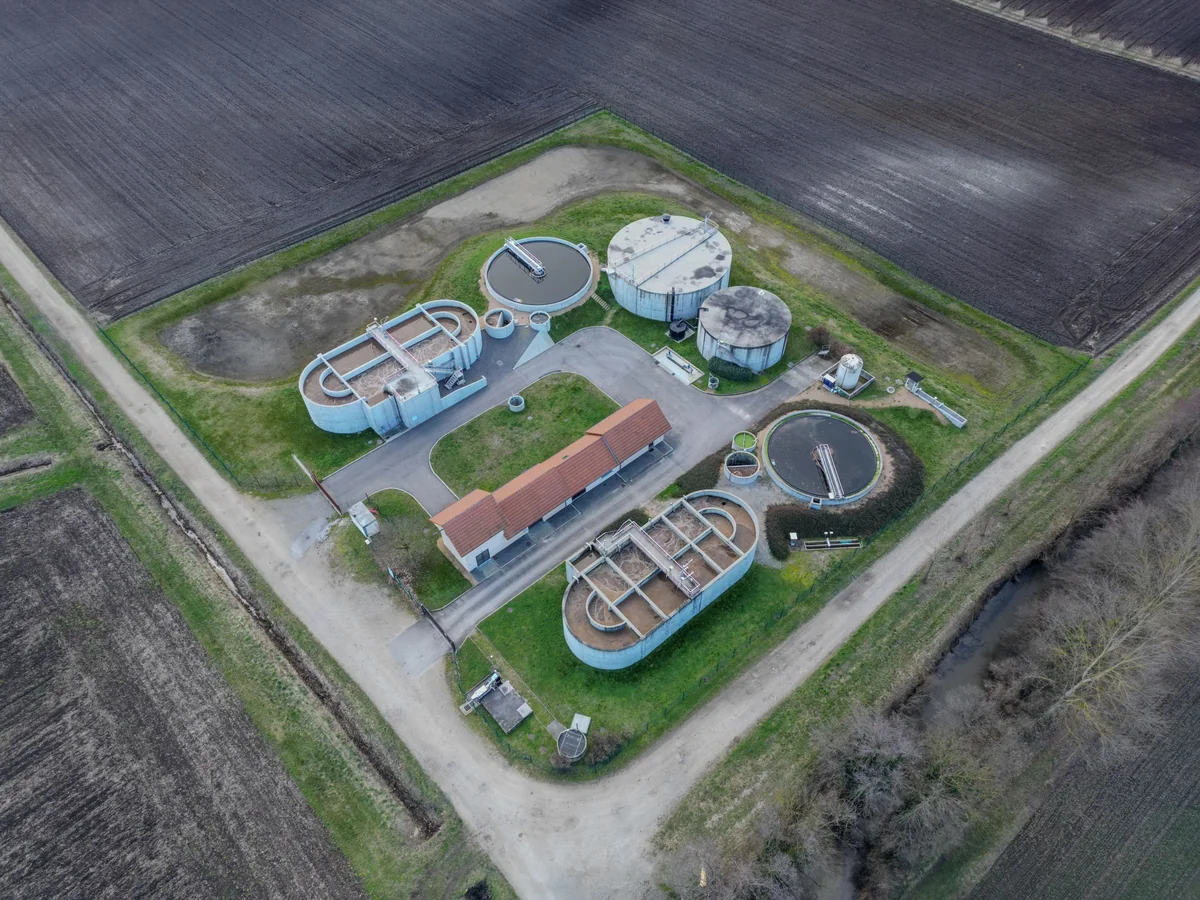 Aerial view of a sewage treatment plant with circular settling tanks, rectangular basins, access roads, cultivated fields and a wooded drainage area.