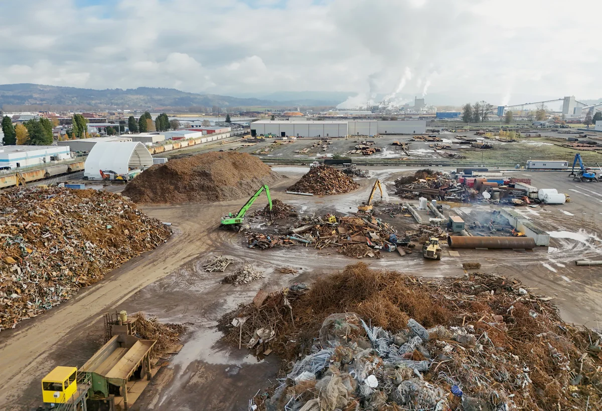 A wide aerial view of a scrap-metal recycling facility with separated piles of metal and wood, excavators, loaders, conveyors, industrial buildings and distant hills under a cloudy sky.