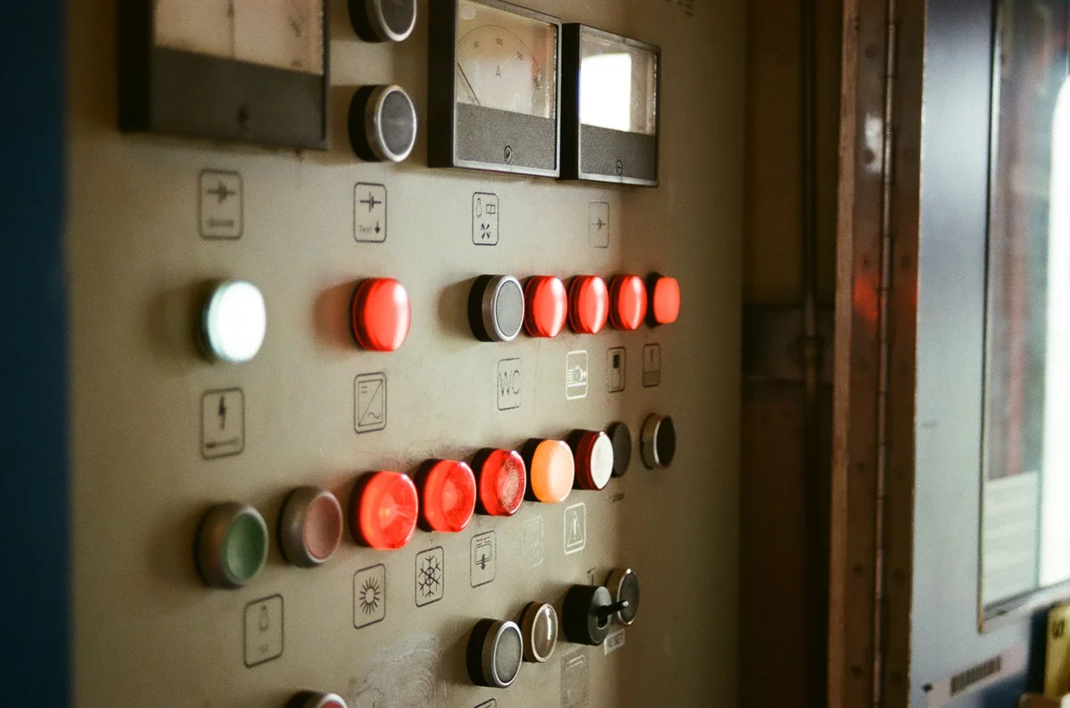 Close-up landscape photograph of an unoccupied industrial control panel with analog gauges, illuminated red and white buttons, switches and instrument markings.