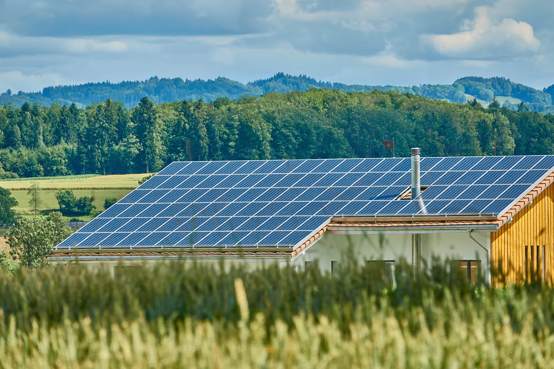 A broad landscape photograph of a building roof covered with rows of blue solar panels, with fields, trees, and wooded hills behind it under a partly cloudy sky.