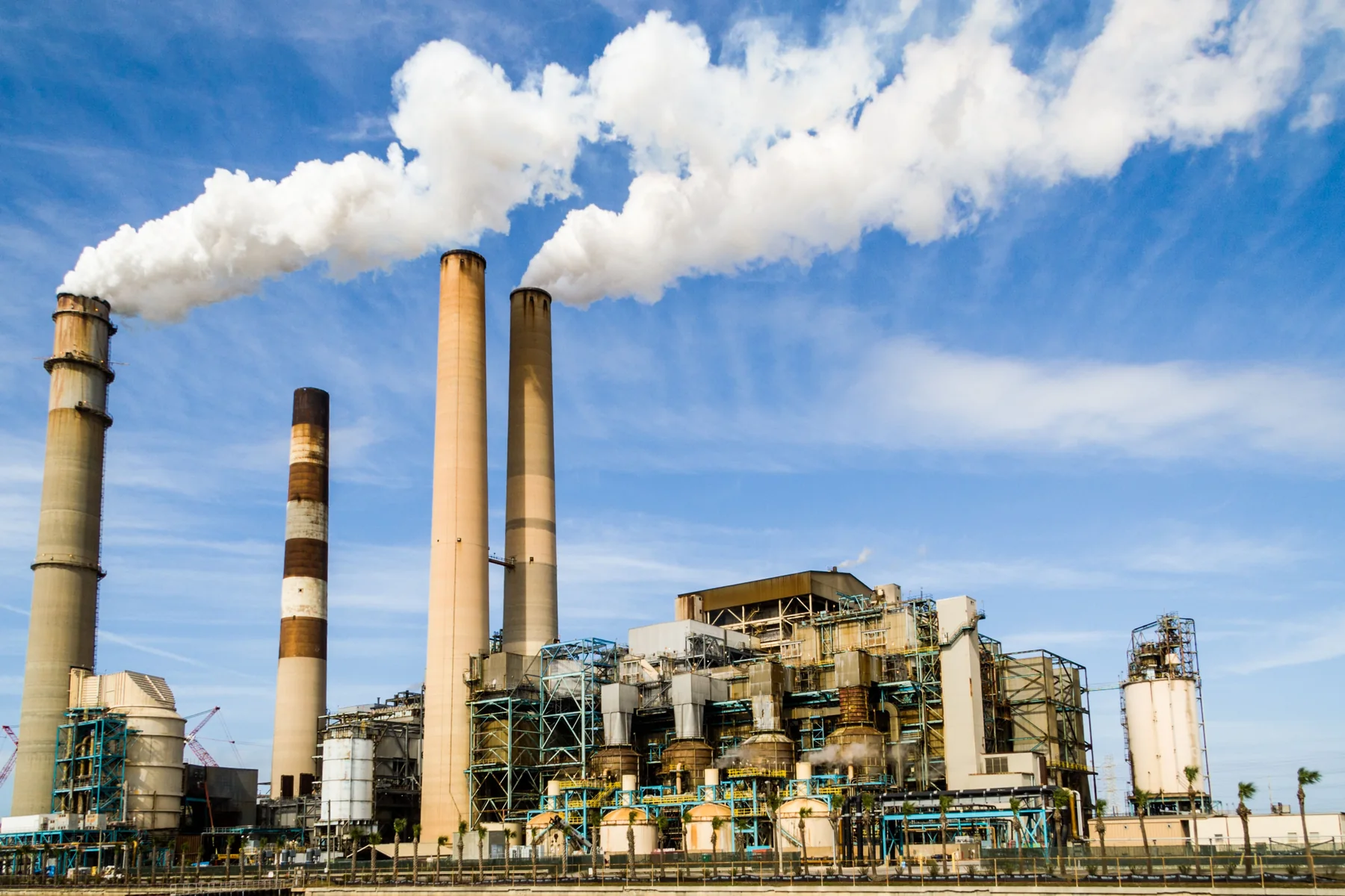 Industrial power plant with several tall smokestacks releasing white plumes above interconnected plant buildings under a blue sky.