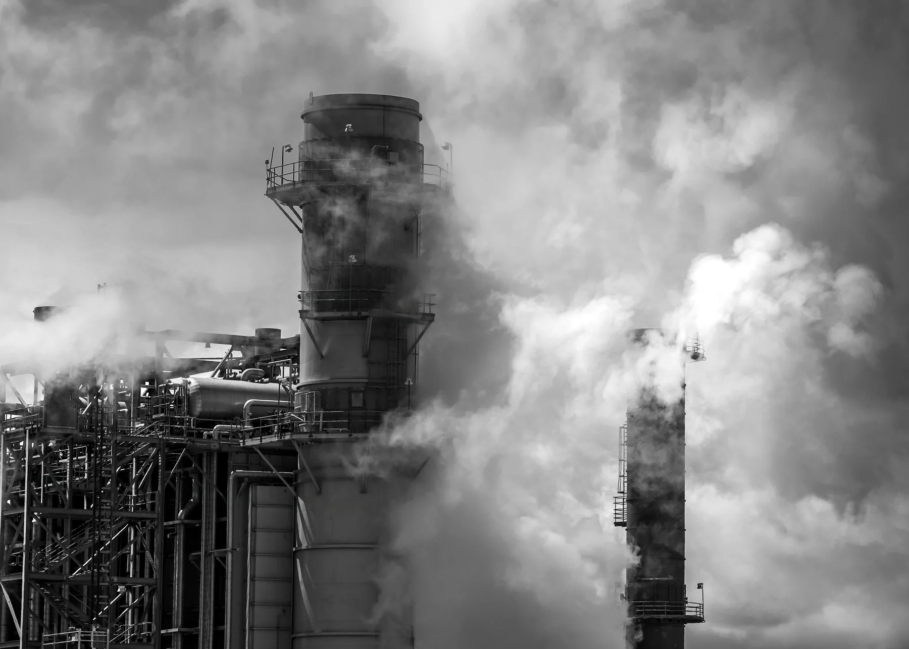 Black-and-white landscape photograph of an industrial processing facility with a tall cylindrical tower, pipework, platforms and thick white vapor or smoke around the structures.