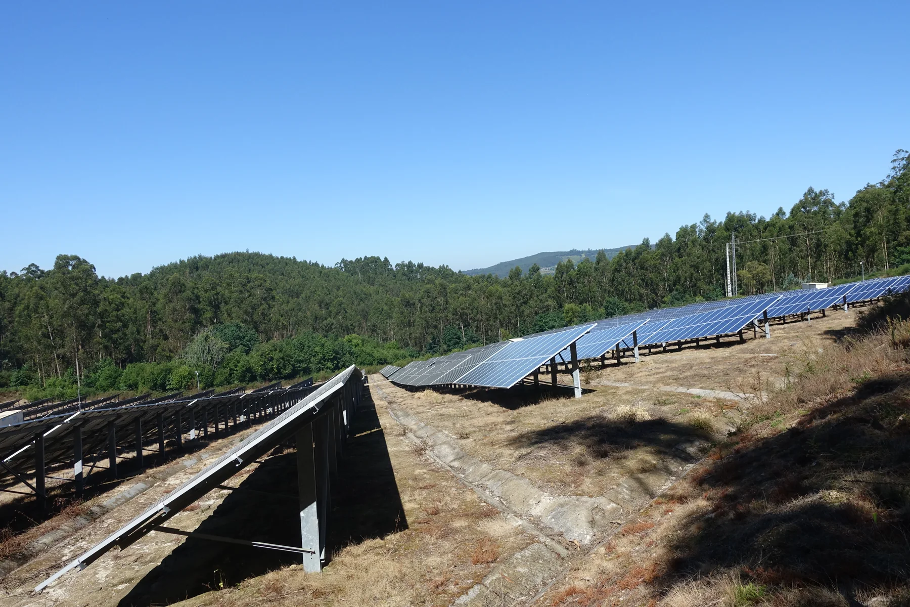 Rows of dark photovoltaic panels form a large solar power station across a green landscape under a cloudy sky.