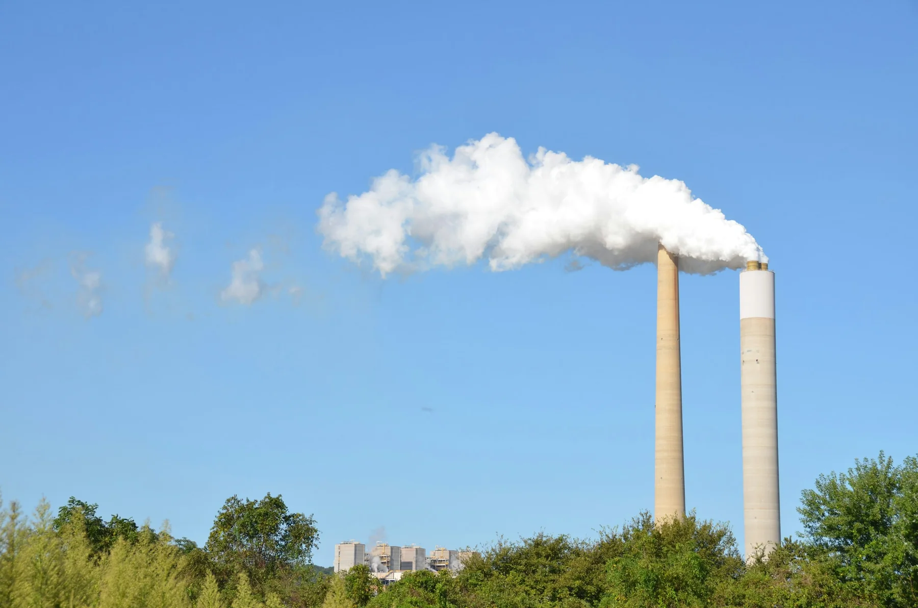Two tall industrial smokestacks release white plumes against a clear blue sky.