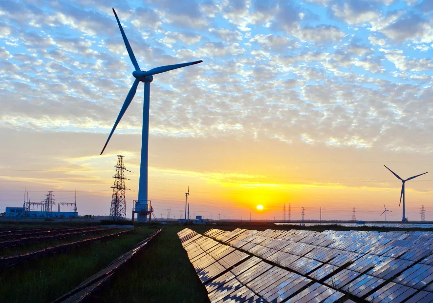 Landscape photograph showing rows of solar panels in the foreground, wind turbines beyond them, and electricity pylons against a low sun.