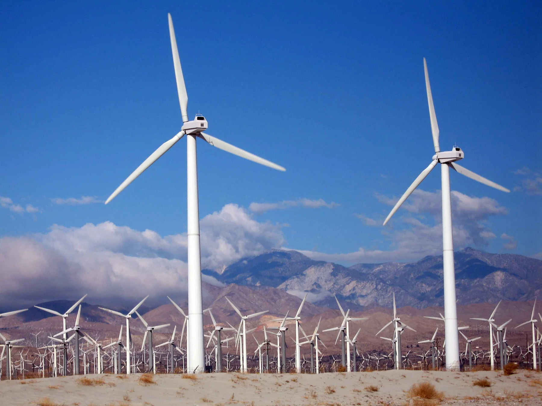 Landscape photograph of multiple white wind turbines spread across a dry valley, with mountain ridges and a blue sky behind them.