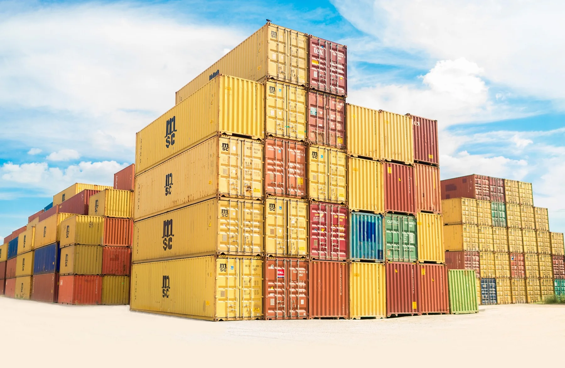 A stack of yellow, red, blue and green intermodal shipping containers stands outdoors beneath a bright blue sky with clouds.