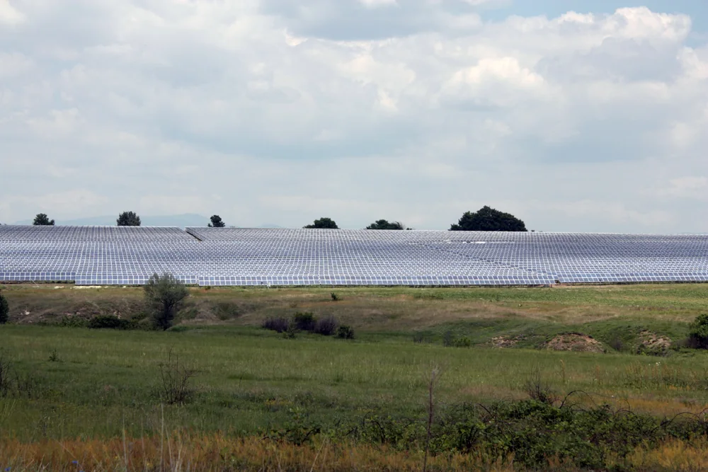 A broad field of densely packed photovoltaic panels stretches across green grassland beneath a cloudy sky, with low hills on the horizon.