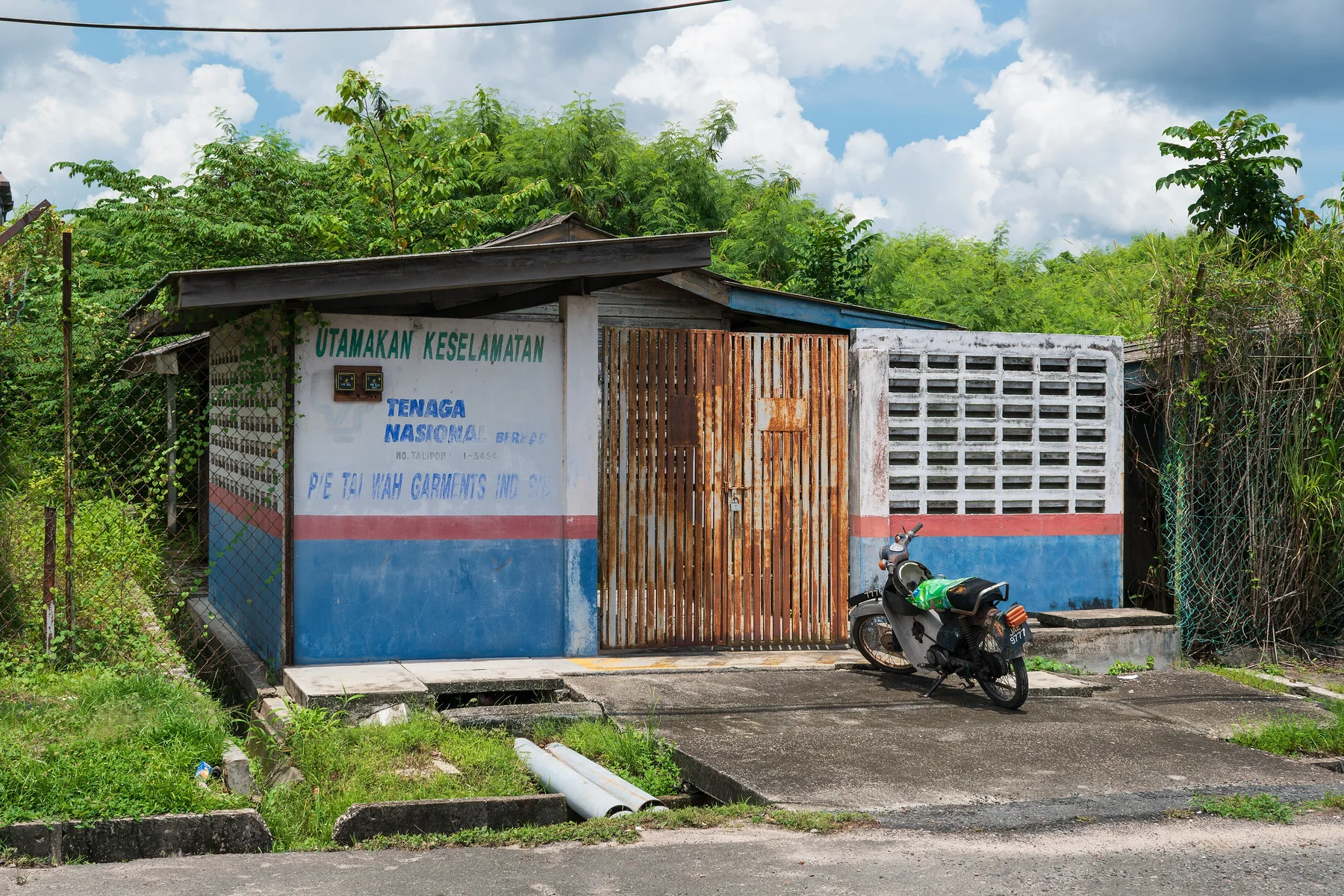 A small Tenaga Nasional electrical substation building beside a road in Kluang, with wall-mounted electrical meters, safety lettering, vegetation, and a parked motorcycle.