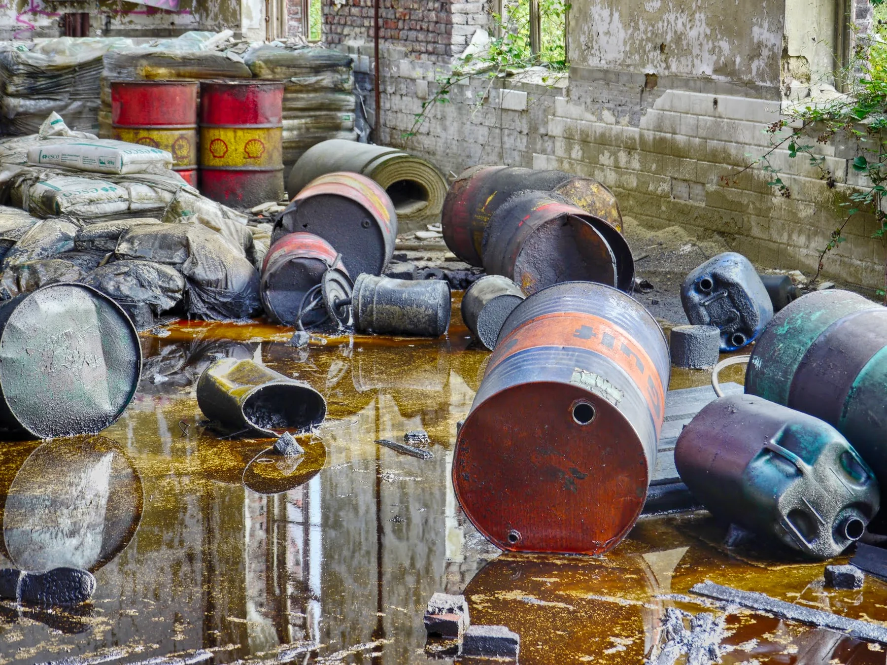 Landscape photograph of assorted rusted and weathered oil drums and containers lying on wet ground beside a weathered industrial masonry wall, with vegetation visible beyond.