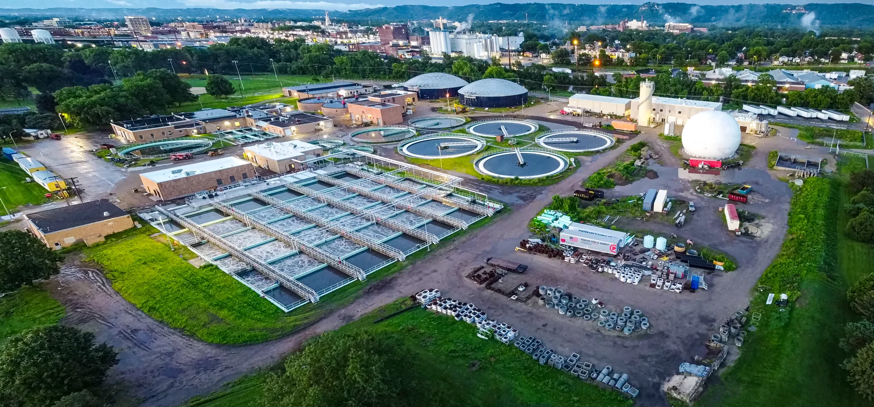 Aerial landscape photograph of a wastewater treatment facility with circular settling tanks, treatment basins, buildings, roads, and surrounding trees.