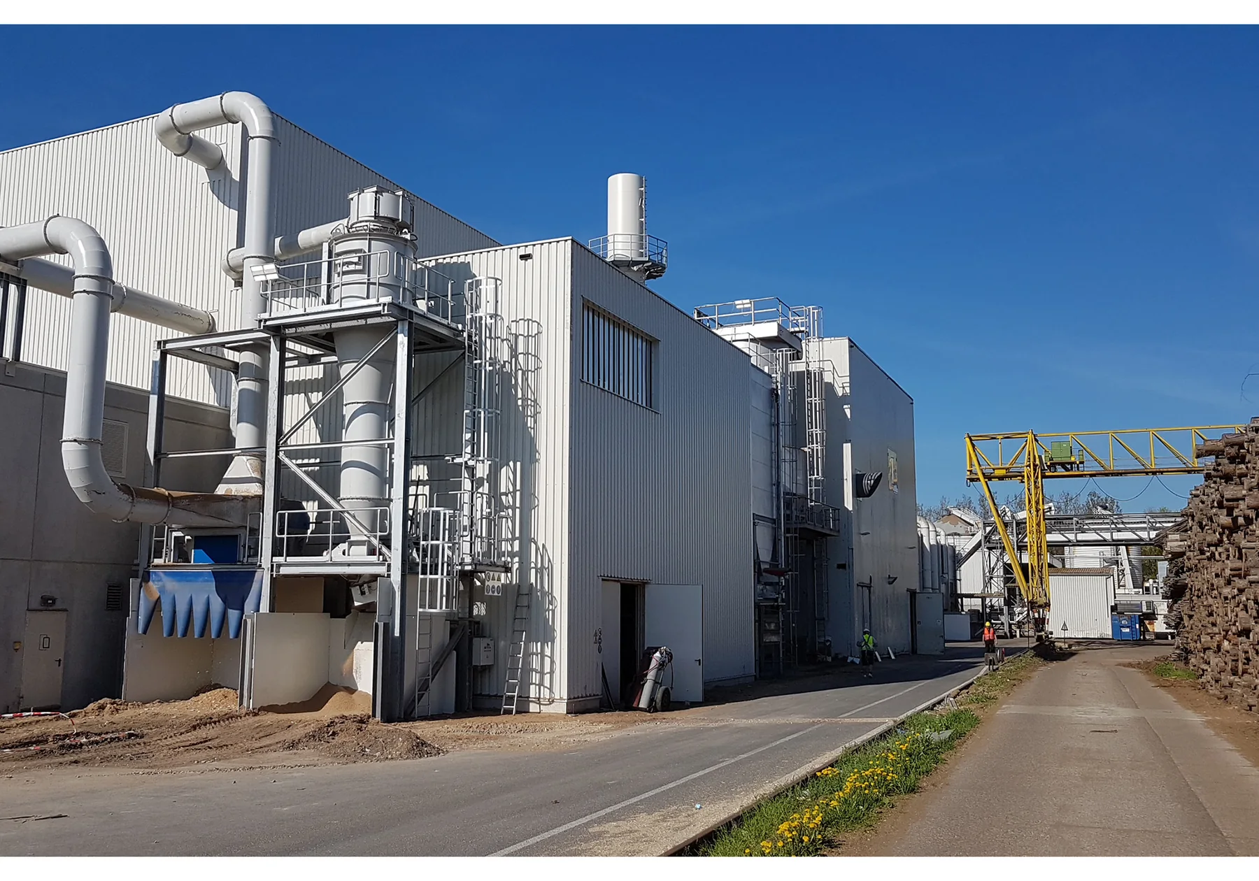 Industrial wood-recycling processing building with ducts, access platforms, a yellow overhead conveyor, and stacked logs at the right under a clear blue sky.