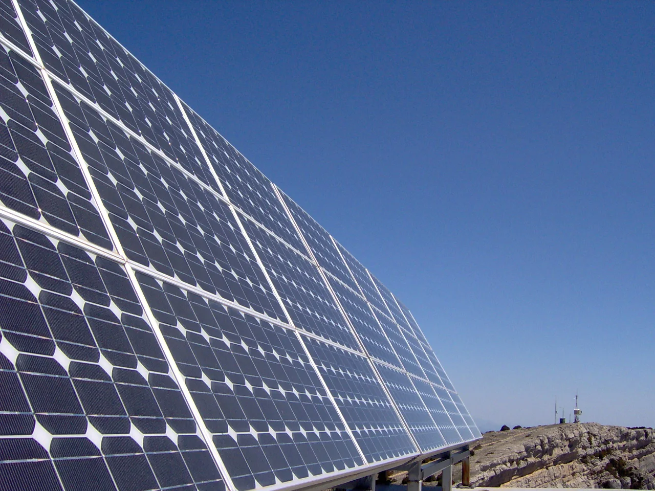 A large array of dark photovoltaic panels rises diagonally across a rocky site beneath a clear blue sky, with a small communications mast in the distance.