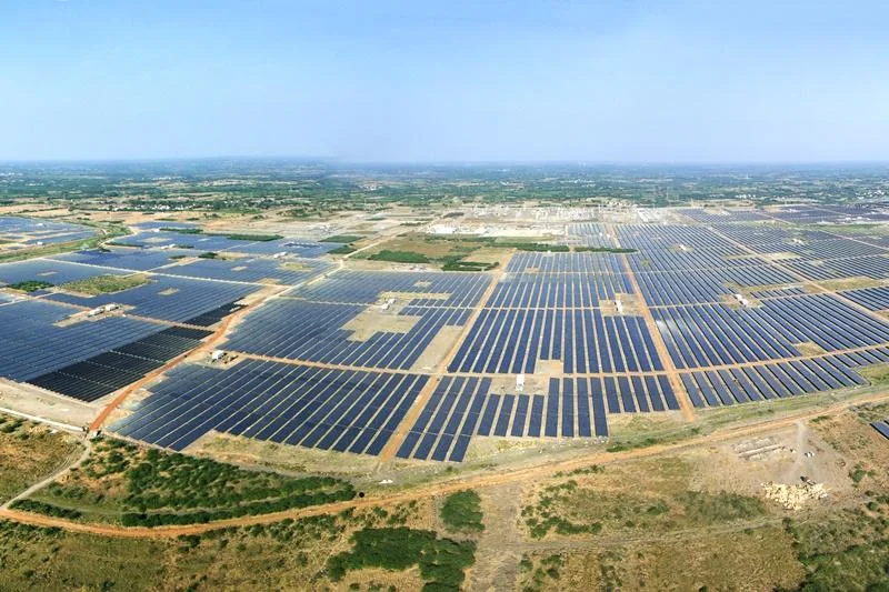 Aerial landscape view of a large solar power park with rows of photovoltaic panels across open ground under a blue sky.